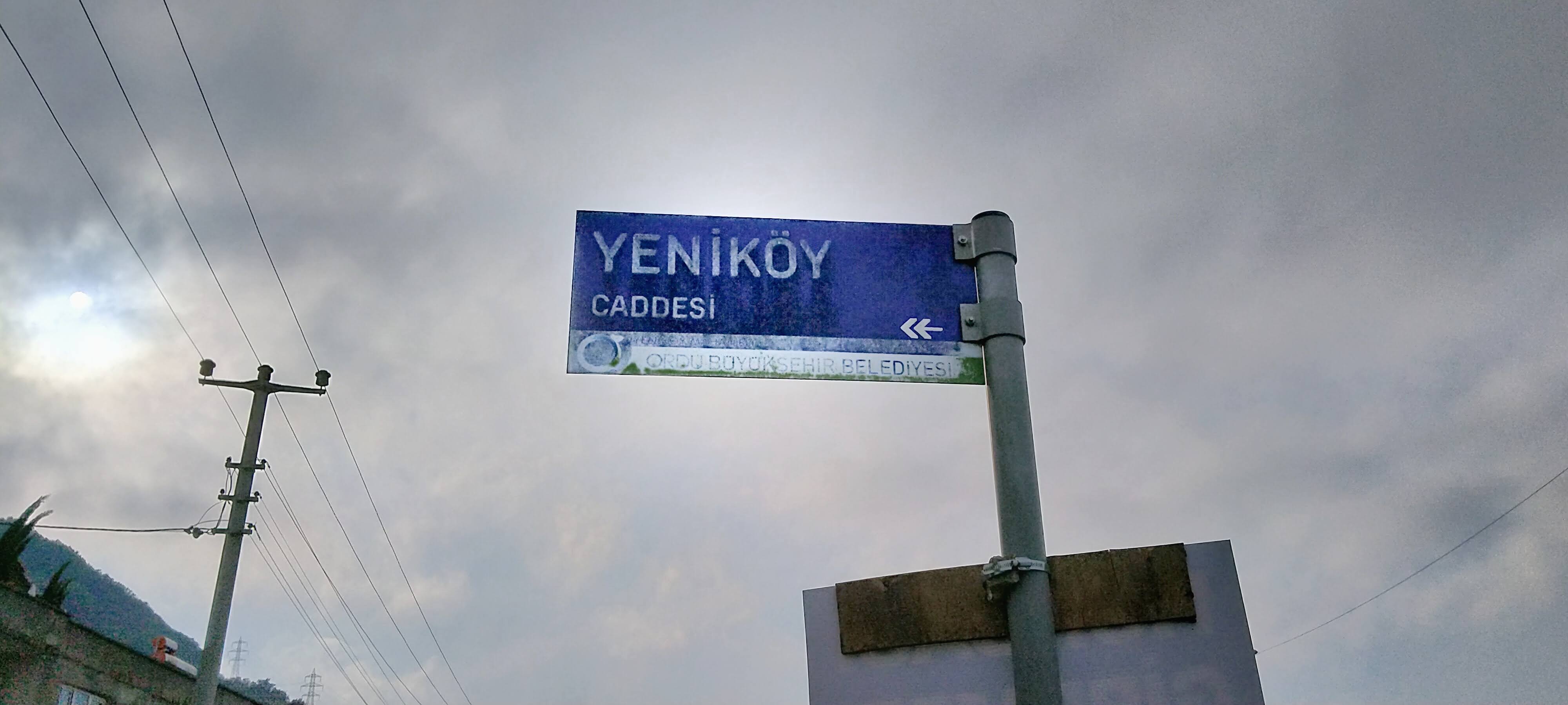Old, fading street sign, with text Yeniköy Caddesi ↞ and the logo of the Metropolitan Municipality of Ordu.